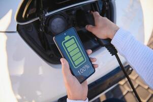 Man Charging Electric Vehicle With Cable Looking At App On Mobile Phone photo