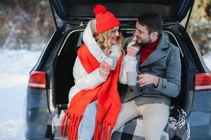 Happy Young couple is sitting in the back trunk of car in winter forest and drinking hot tea from thermos. Winter time photo