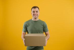 Delivery man in green uniform isolated on yellow background, studio portrait. Male employee working as courier dealer hold empty cardboard box. Service concept. Mock up copy space photo