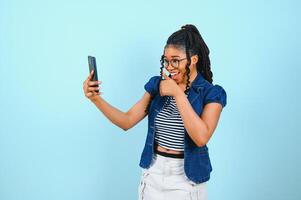 Happy cheerful Afro American woman looks at screen of smart phone enjoys online chatting types text message surfs social networks dressed casually poses against blue background. Technology concept. photo