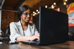 Beautiful african business woman waiting for the meeting in a co-working space and sitting with the portable computer. Hipster girl is surfing the web on a laptop during lunch. photo