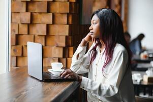 Beautiful african business woman waiting for the meeting in a co-working space and sitting with the portable computer. Hipster girl is surfing the web on a laptop during lunch. photo