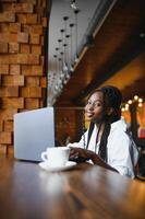Beautiful african business woman waiting for the meeting in a co-working space and sitting with the portable computer. Hipster girl is surfing the web on a laptop during lunch. photo