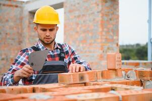 Using bricks. Young construction worker in uniform is busy at the unfinished building photo
