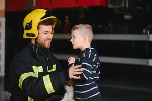 A firefighter take a little child boy to save him. Fire engine car on background. Fireman with kid in his arms. Protection concept. photo