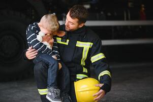 Dirty firefighter in uniform holding little saved boy standing on black background. photo