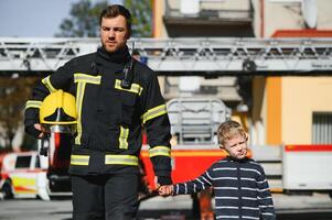 A firefighter take a little child boy to save him. Fire engine car on background. Fireman with kid in his arms. Protection concept. photo
