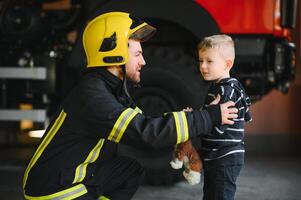 A firefighter take a little child boy to save him. Fire engine car on background. Fireman with kid in his arms. Protection concept. photo