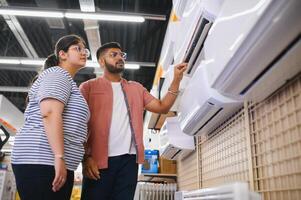 Young indian couple, satisfied customers choosing air conditioner in appliances store photo