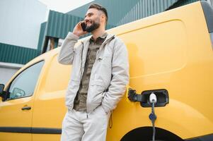 bearded caucasian man standing near an electric car that is charging and making time adjustments on a smartphone. photo
