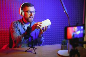 Young male technology blogger recording blog or vlog about new smartphone and other gadgets at home studio. Blogging, Work from Home concept. Focus on gadgets at the table. Web Banner photo