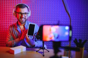 Young male technology blogger recording blog or vlog about new smartphone and other gadgets at home studio. Blogging, Work from Home concept. Focus on gadgets at the table. Web Banner photo