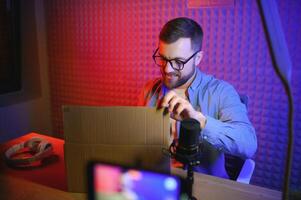 Panoramic view of young adult vlogger creating internet content, holding carton box in hands, unpacking received pack and recording media online on broadcast equipment photo