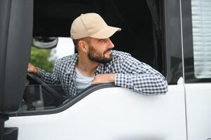 A driver sitting inside the semi-truck while looking through the open window with multiple trucks parked in the background photo