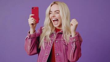 Joyful Young Woman Celebrating Success While Holding Red Smartphone Against Purple Background photo