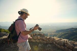 Man with hat and backpack taking photos of scenic mountain landscape on a sunny day