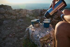 Person pouring coffee from thermos into cups during outdoor adventure, with rocky landscape in background photo