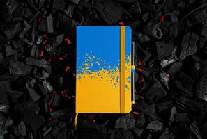 A small closed notebook with a yellow-blue cover in the colors of the Ukrainian flag. Texture of charcoal. Dark back photo