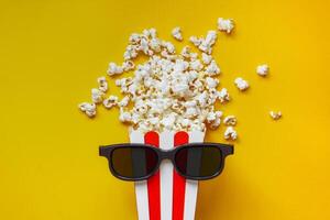 Popcorn in a red and white cardboard box with glasses for a 3D movie on the yellow back. Popcorn in red striped bucket on yellow background. Flat lay concept. photo