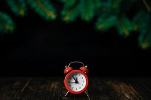 Red alarm clock on a dark wooden background with fir branches. Five to twelve on the clock. Christmas card, blank. photo