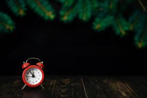 Red alarm clock on a dark wooden background with fir branches. Five to twelve on the clock. Christmas card, blank. photo