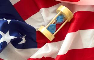 Hourglass on table with USA flag in the background. Sand glass on old wooden table with American flag in the background, vertical image. photo