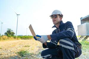 A technician in safety gear meticulously records data with a wind turbine in the backdrop at dusk. photo