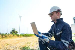 A technician in safety gear meticulously records data with a wind turbine in the backdrop at dusk. photo