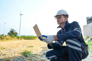 A technician in safety gear meticulously records data with a wind turbine in the backdrop at dusk. photo