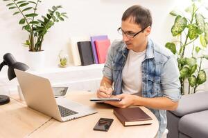 Happy young man, wearing glasses and smiling, as he works on his laptop to get all his business done early in the morning with his cup of coffee photo