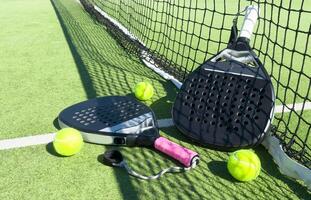 Yellow balls on grass turf near padel tennis racket behind net in green court outdoors with natural lighting. Paddle is a racquet game. Professional sport concept with copy space. photo
