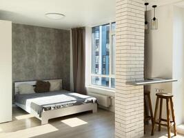 Beautiful modern bedroom with different walls and a parquet on the floor. There is a bed with light pillows and blanket, big windows with a glass door to the terrace with wicker chairs, TV, stand. photo
