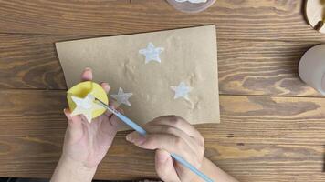 Woman applies gouache paint with brush to potato stamp in form of star and makes white print on craft paper. Hands decorate scrapbooking wrapping paper on wooden table. Diy and handmade on quarantine. video