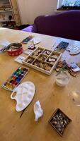 Large desk with watercolors and gouache paints brushes and palette and box with plywood badges. Drawing for children. Cozy workshop with purple sofa and wooden table. video