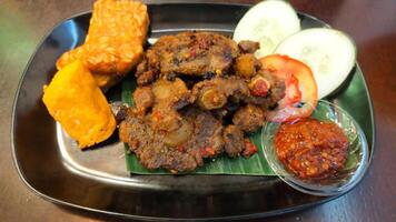 Spicy Grilled Oxtail with Tofu and Tempeh Served with Fresh Cucumber and Tomato Slices and Spicy Chili Sauce on Black Plate photo