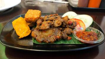 Spicy Grilled Oxtail with Tofu and Tempeh Served with Fresh Cucumber and Tomato Slices and Spicy Chili Sauce on Black Plate photo