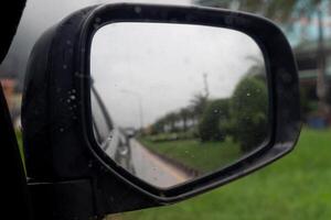 Inside view of mirrors wing rear view of a gray car with drop of water rain on the glass. Driving on aspahlt road beside with green grass. Under of white clouds of rainy. photo