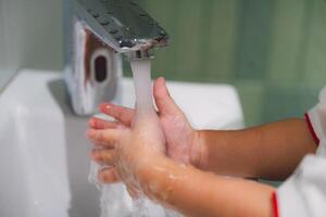 Washing Hands. A close-up shot of a child's hands diligently washing under running water in a bathroom sink. The image emphasizes the importance of hygiene and cleanliness, with a focus on the gentle photo
