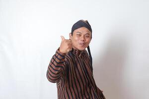 Portrait of amazed Asian man in javanese cloth with blangkon hat showing approval and agreement sign, making thumbs up hand gesture. Isolated image on white background photo