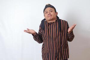 Portrait of confused Asian man in javanese cloth with blangkon hat pointing sideways, making choice, choosing between two objects. Isolated image on white background photo