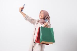 Portrait of excited Asian hijab woman in casual suit holding credit card and shopping bags while taking selfie with mobile phone. Shopaholic girl concept. Isolated image on white background photo