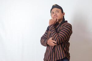 Portrait of curious Asian man in javanese cloth with blangkon hat thinking hard and feeling confused. Isolated image on white background photo