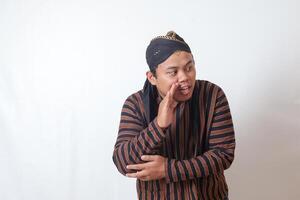 Portrait of serious Asian man in javanese cloth with blangkon hat whispering malicious talk conversation, hand on mouth telling secret rumor. Isolated image on white background photo