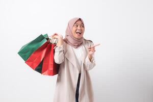 Portrait of excited Asian hijab woman in casual suit holding paper shopping bags while pointing with finger. Shopaholic girl and discount buying concept. Isolated image on white background photo