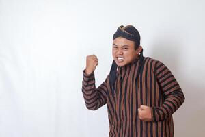 Portrait of excited Asian man in javanese cloth with blangkon hat raising his fist, celebrating success after winning a competition. Isolated image on white background photo