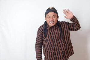 Portrait of friendly Asian man in javanese cloth with blangkon hat saying hi, waving hand at camera, greeting person with happy smile. Isolated image on white background photo