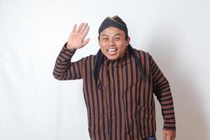 Portrait of friendly Asian man in javanese cloth with blangkon hat saying hi, waving hand at camera, greeting person with happy smile. Isolated image on white background photo