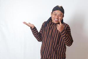 Portrait of excited Asian man in javanese cloth with blangkon hat showing product and pointing with hand and finger to the side. Sales and promotion concept. Isolated image on white background photo