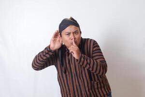 Portrait of serious Asian man in javanese cloth with blangkon hat whispering malicious talk conversation, hand on mouth telling secret rumor. Isolated image on white background photo