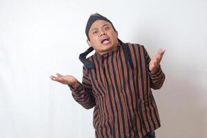 Portrait of confused Asian man in javanese cloth with blangkon hat pointing sideways, making choice, choosing between two objects. Isolated image on white background photo